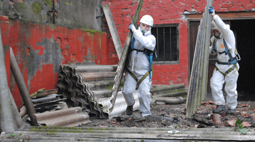 Kosten Asbestentsorgung: Zwei Handwerker in Schutzausrüstung tragen zwei Asbestplatten