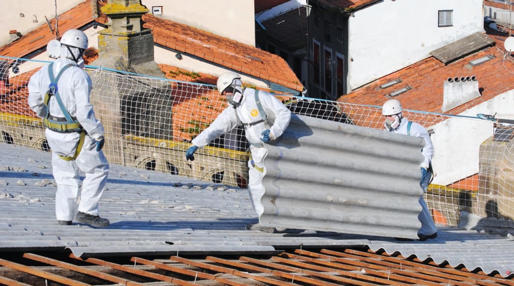 Drei Handwerker in Voll-Schutzausrüstung entfernen alte Asbestplatten vom Dach.